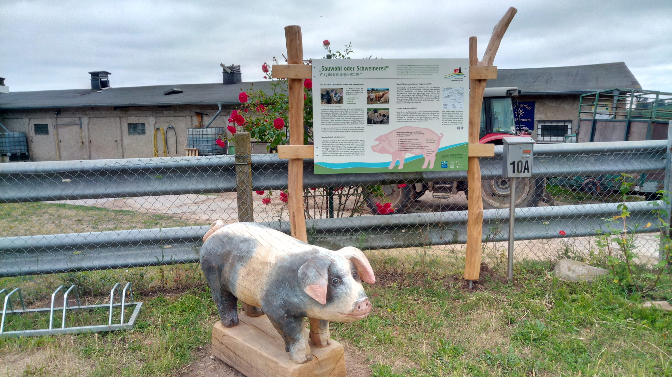 Stallgebäude mit Zaun und Infotafel, davor eine Holzskulptur in Form eines Schweins.