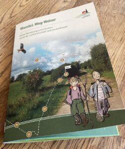 ein Heft auf einem Tisch, Titel WandeLwegweiser, mit Foto eines Feldwegs und Zeichnung mit wandernden Kindern